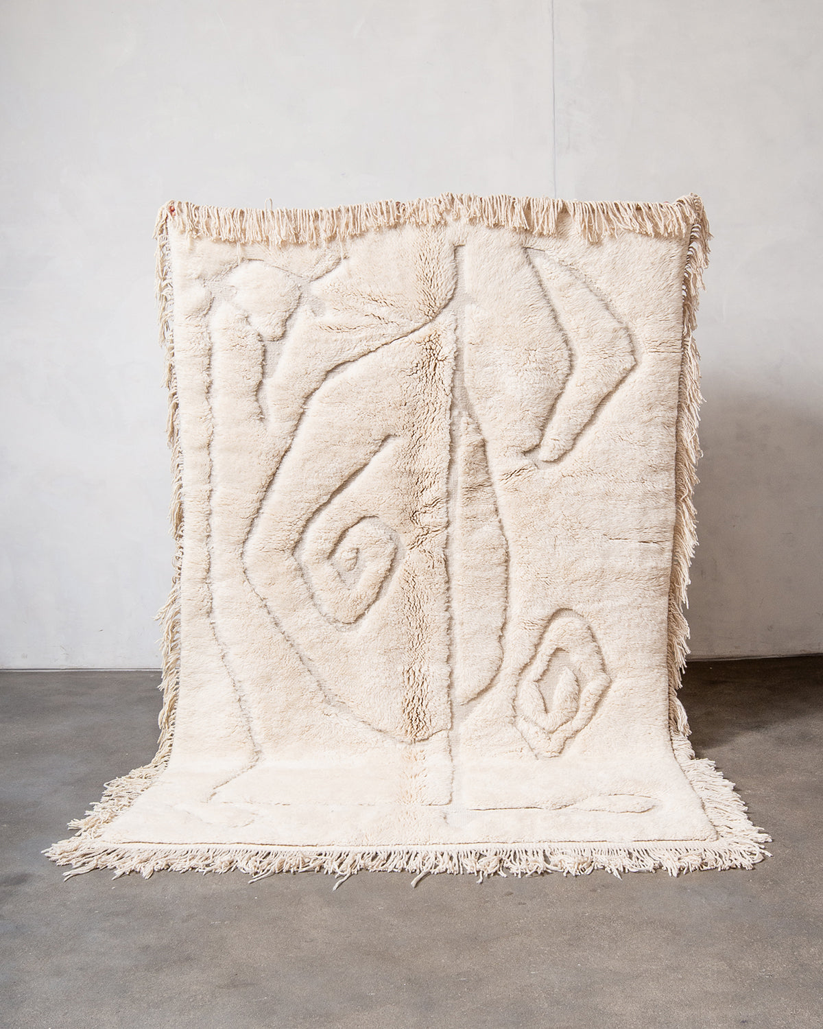 Berberlin x Malaika Raiss Beni M'rirt rug made of velvety cream sheep's wool with natural wool tassels on all four sides, being held up against a white wall and concrete floor.