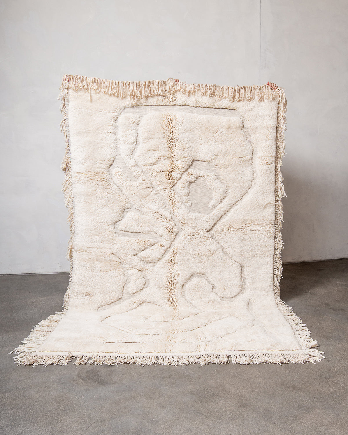 Berberlin x Malaika Raiss Beni M'rirt rug made of velvety cream sheep's wool with natural wool tassels on all four sides, being held up against a white wall and concrete floor.
