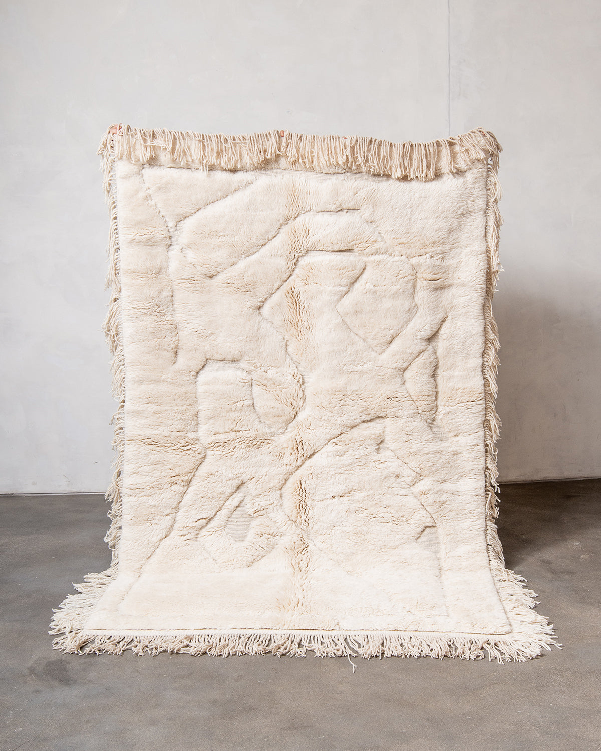 Berberlin x Malaika Raiss Beni M'rirt rug made of velvety cream sheep's wool with natural wool tassels on all four sides, being held up against a white wall and concrete floor.