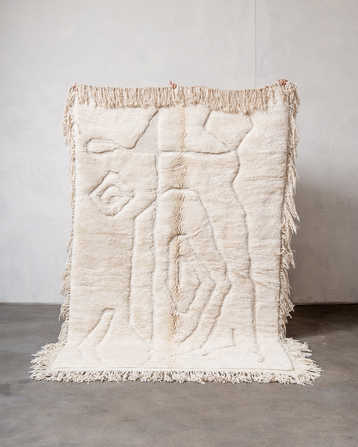 Berberlin x Malaika Raiss Beni M'rirt rug made of velvety cream sheep's wool with abstract woven design and natural wool tassels on all four sides, being held up by hands against white wall and concrete floor.