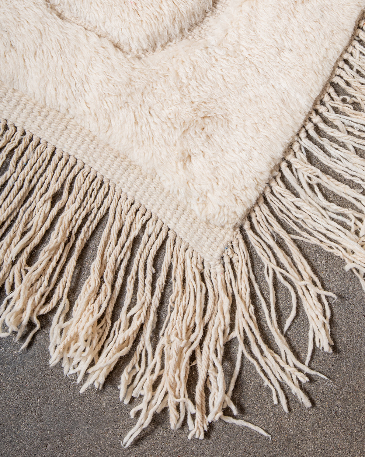 Corner of Beni M'rirt rug made of thick cream sheep's wool with long natural wool tassels on both sides, laying on concrete studio floor.