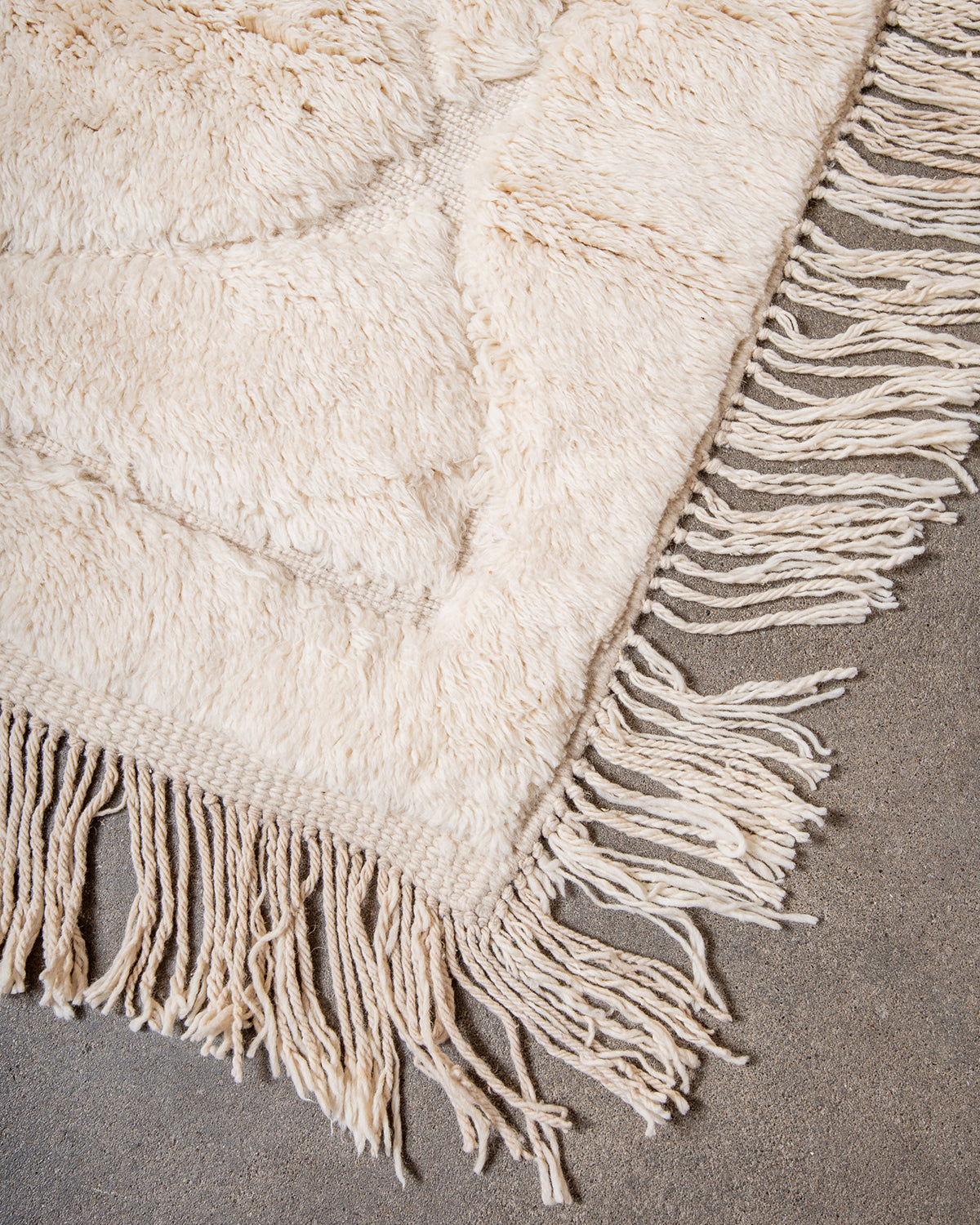 Corner of Beni M'rirt rug made of thick cream sheep's wool with long natural wool tassels on both sides, laying on concrete studio floor.