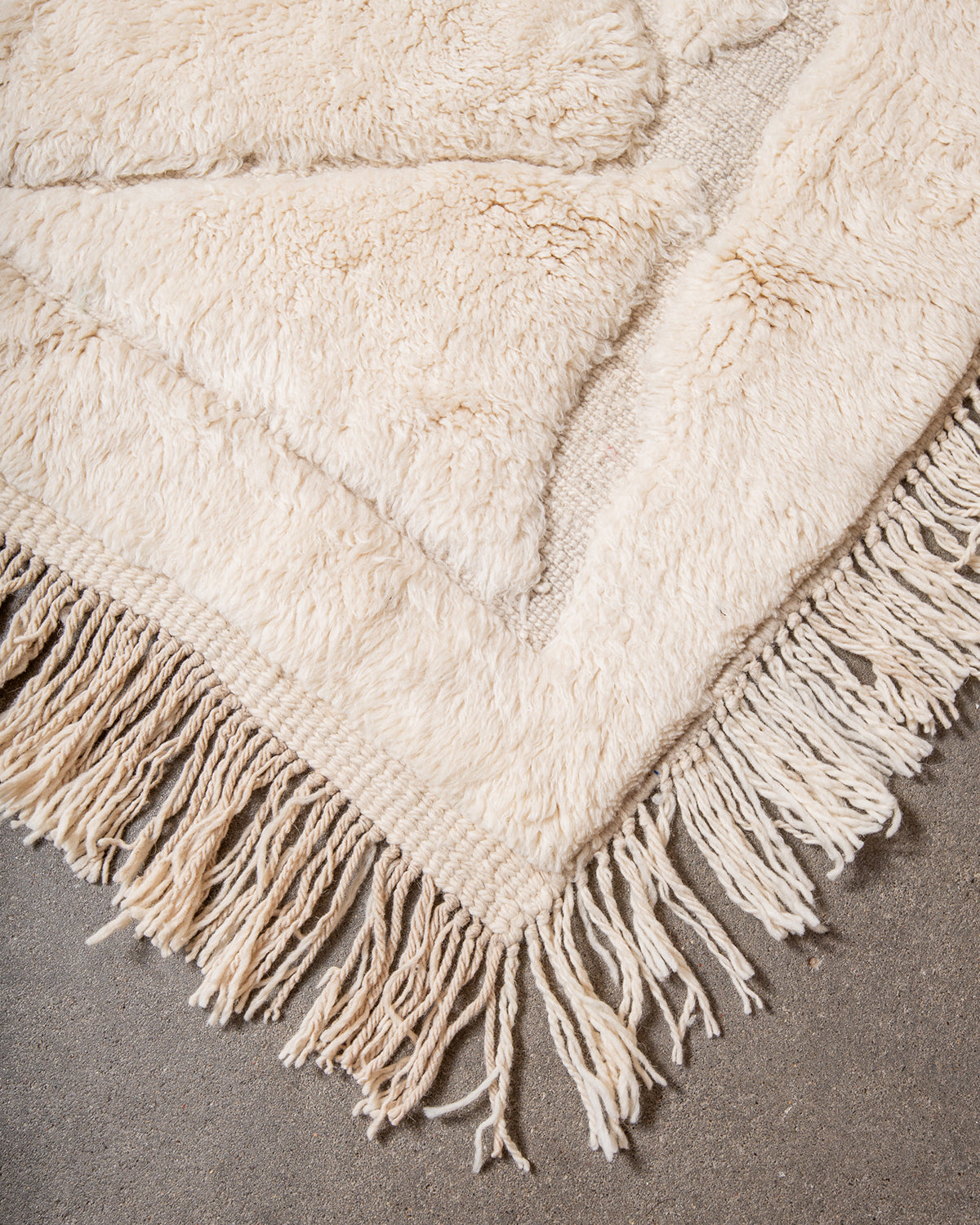 Corner of Beni M'rirt rug made of thick cream sheep's wool with long natural wool tassels on both sides, laying on concrete studio floor.