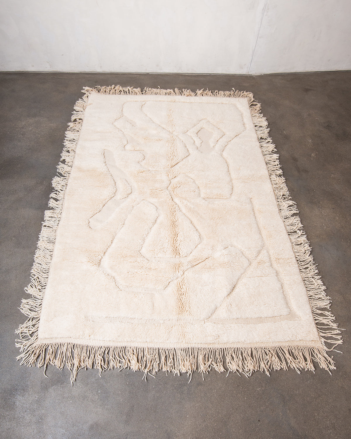 Berberlin x Malaika Raiss Beni M'rirt rug made of velvety cream sheep's wool with abstract woven design and natural wool tassels on all four sides, laying on concrete studio floor.