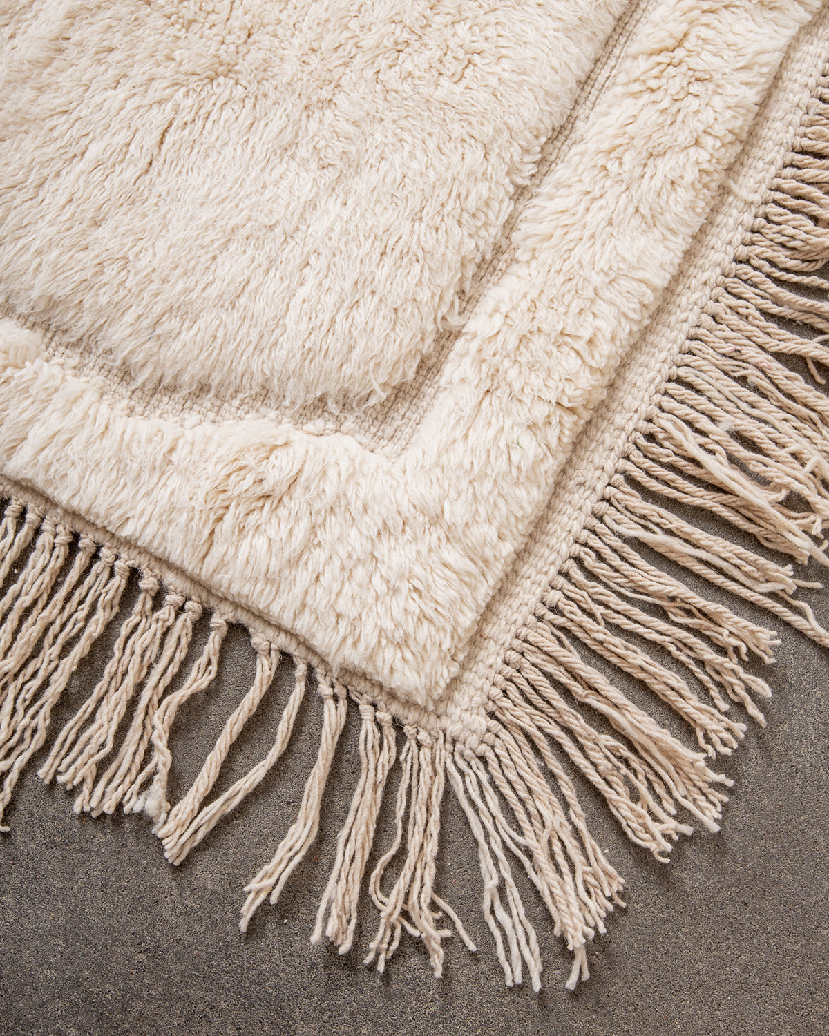 Corner of Beni M'rirt rug made of thick cream sheep's wool with long natural wool tassels on both sides, laying on concrete studio floor.