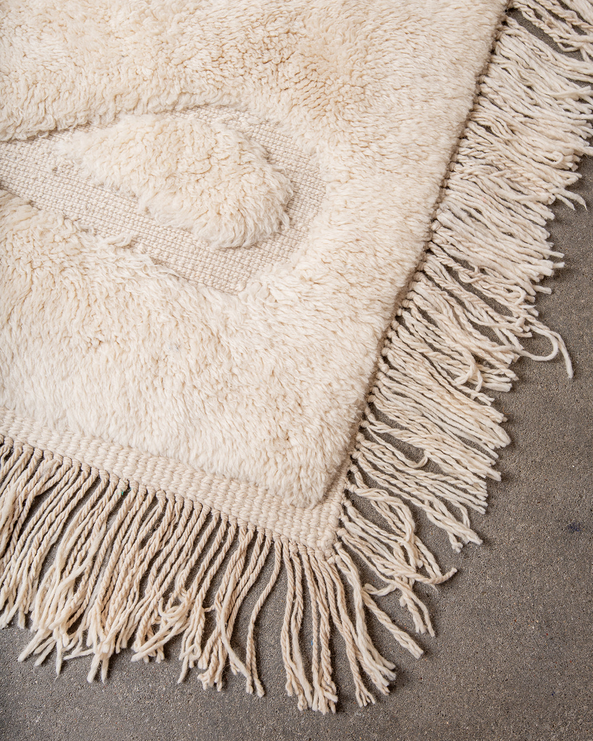 Corner of Beni M'rirt rug made of thick cream sheep's wool with long natural wool tassels on both sides, laying on concrete studio floor.