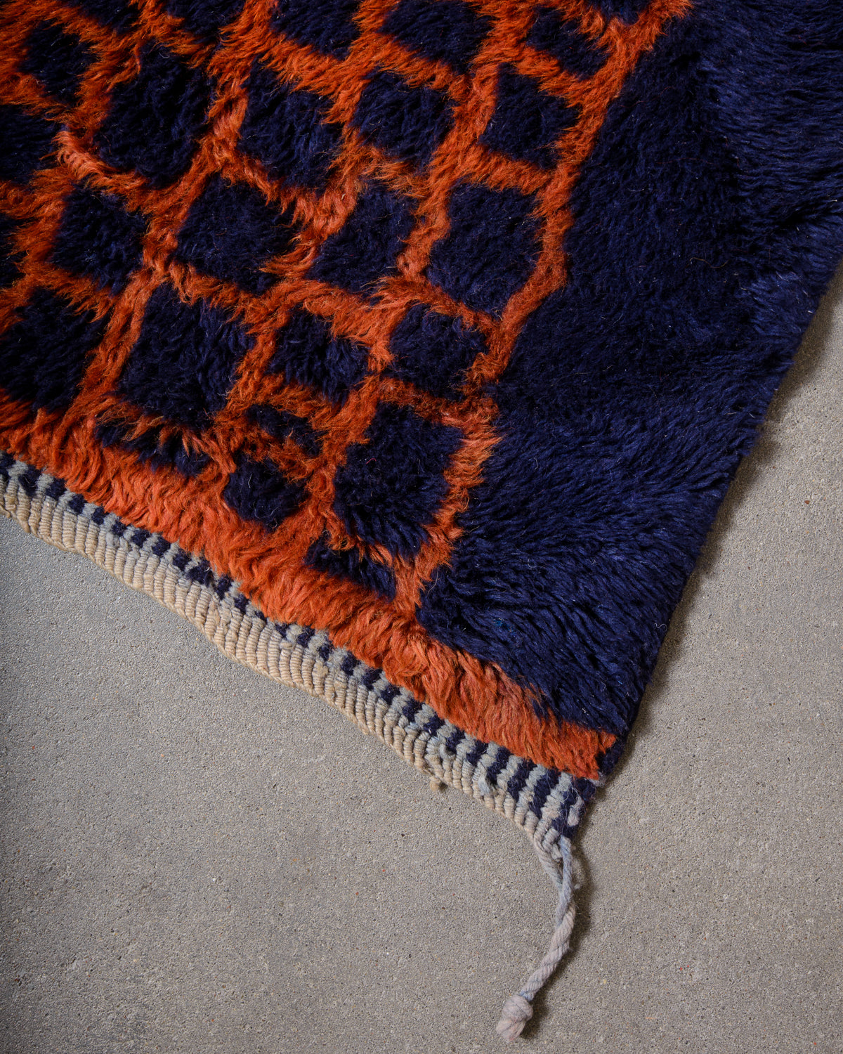 Corner of wool rug laying on concrete studio floor with tightly woven edge in alternating light blue and dark blue and finished with natural cream wool and single light blue tassel.