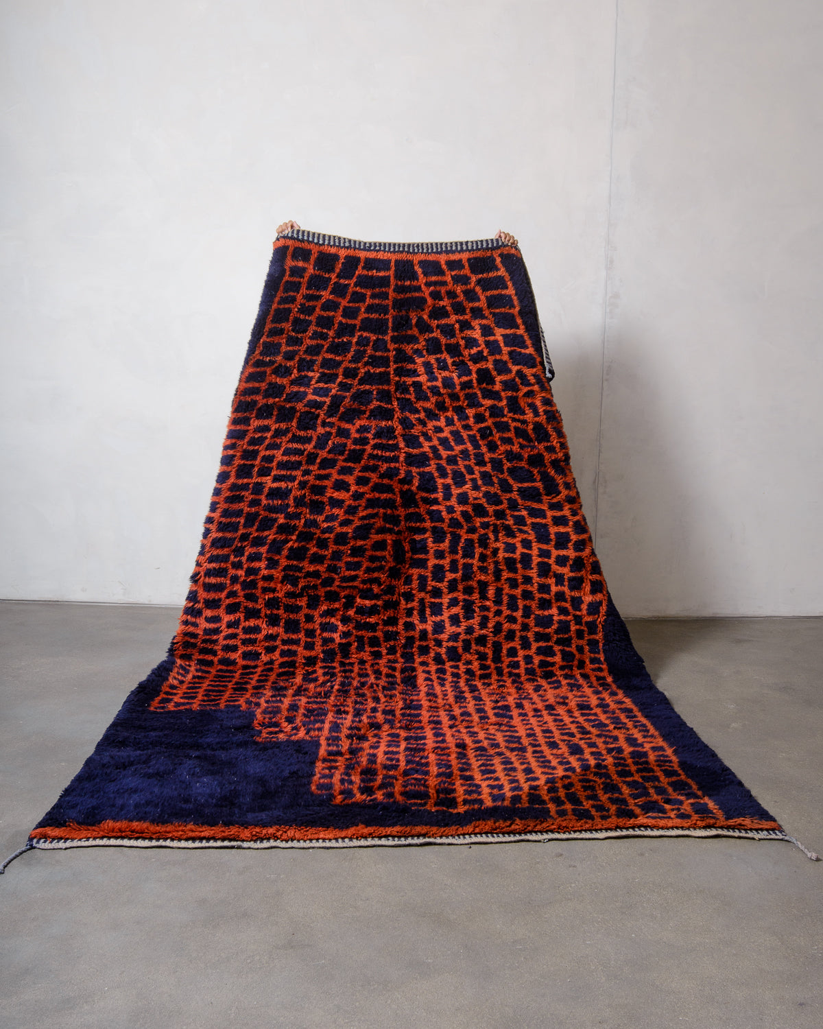 Berberlin Beni M'rirt rug in deep navy blue with contrasting rust red abstract grid pattern held up by two hands against white wall and grey floor.