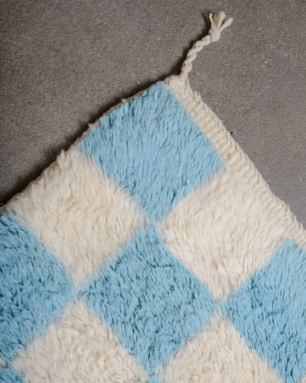 Corner tassel and tightly woven edge of Beni M'rirt wool rug in white and light blue checkerboard design, laying flat on a concrete floor. 