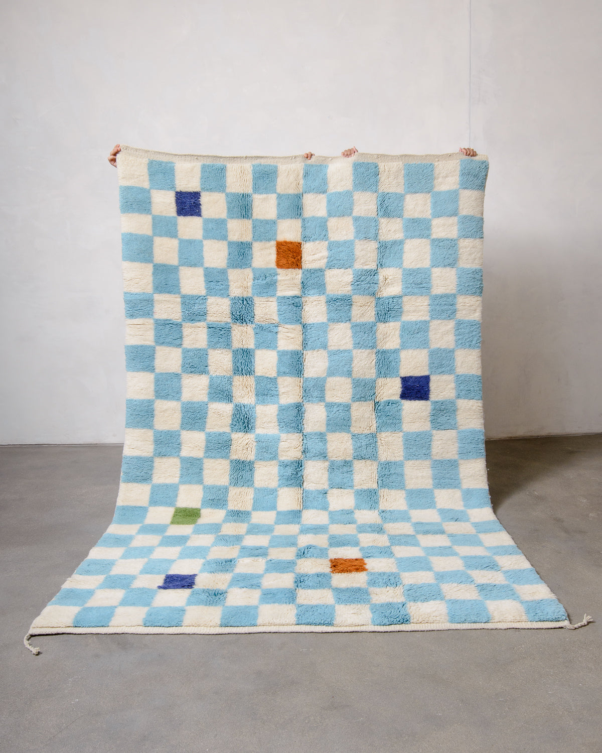 Berberlin Beni M'rirt wool rug in cream and light blue checkerboard design, held up against a white wall and grey floor. Some of the checkered squares are a different colour. 