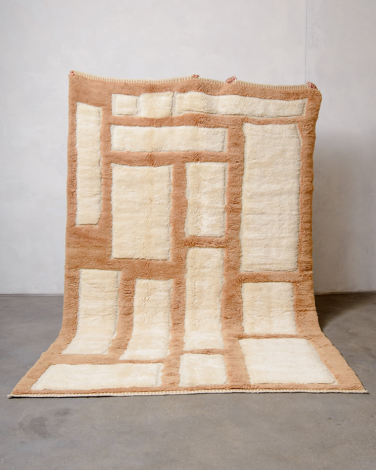 Berberlin Beni M'rirt wool rug in cream and light brown geometric design, being held up against white wall and grey cement floor.