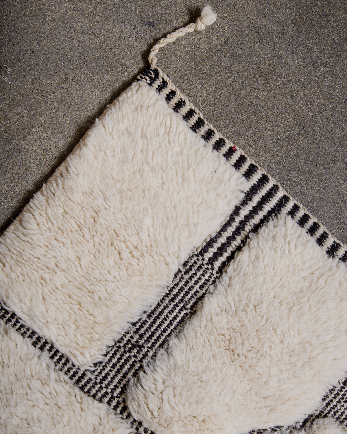 Corner of Beni M'rirt rug in cream and black wool with woven edges and single tassel on cement floor.. 