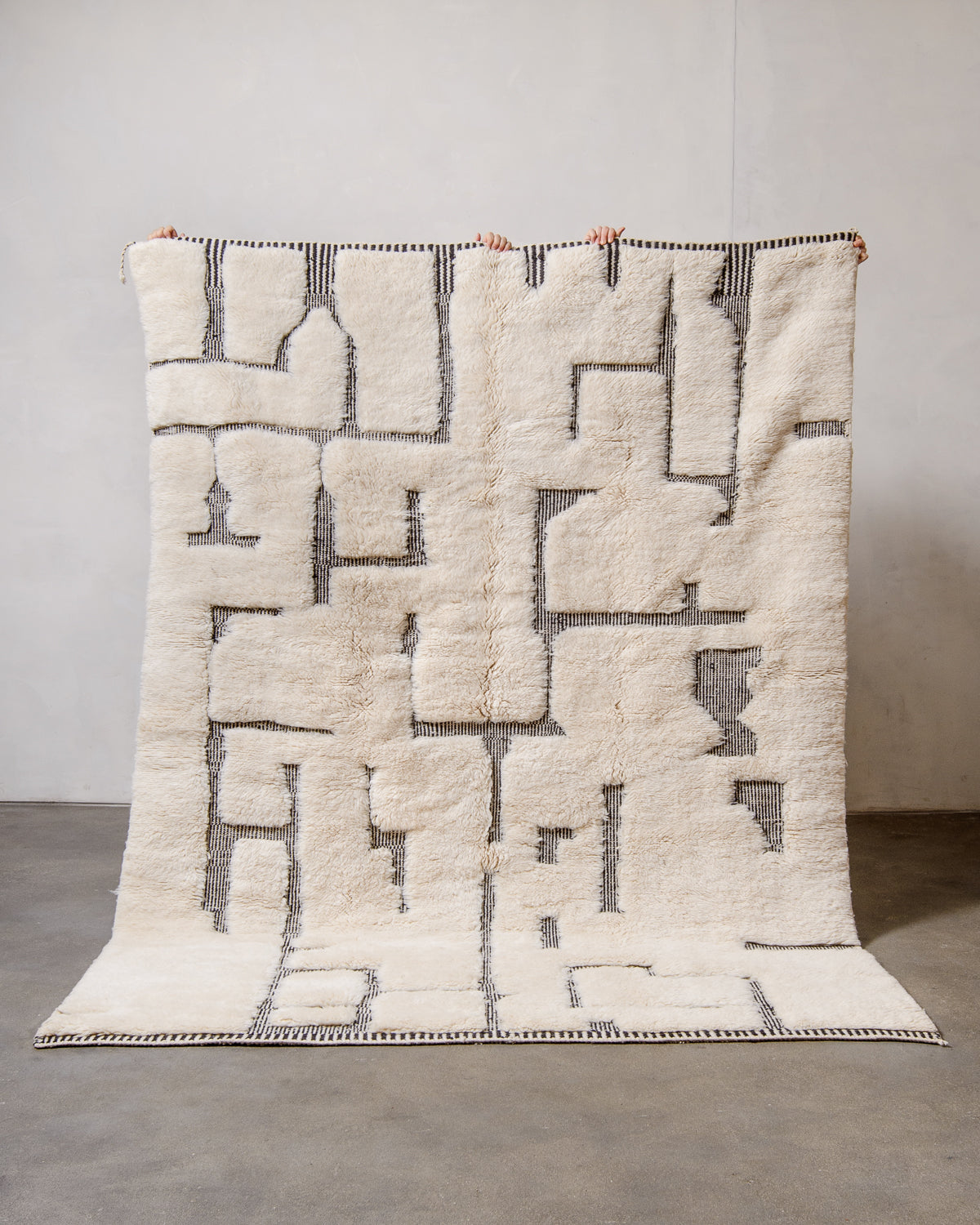 Berberlin Beni M'rirt rug in cream wool with flat-woven design details in thin black and white stripes, held up against a white wall and grey floor.