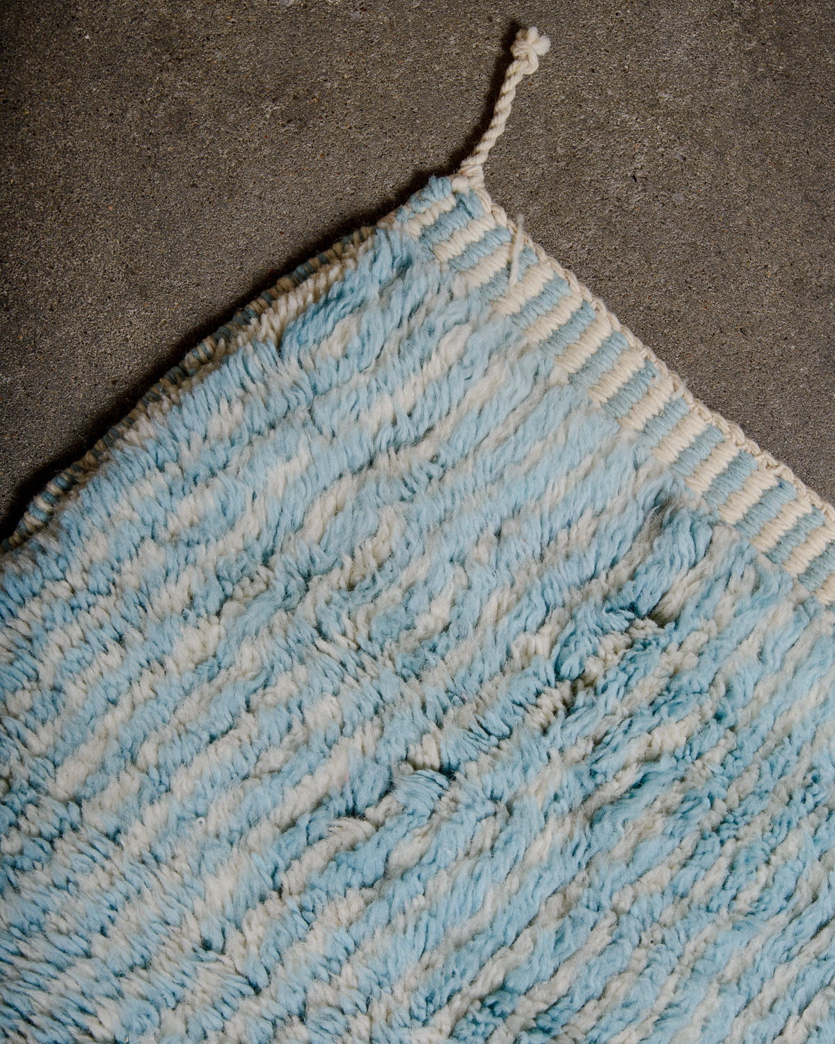 Corner of Beni M'rirt rug in cream and light blue stripes showing the woven edge and single corner tassel.