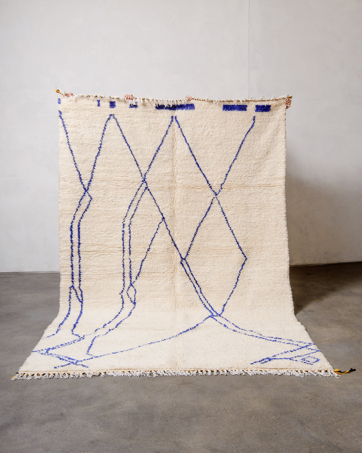 Wool Azilal rug in cream with a blue abstract design being held up by hands against a white wall and concrete floor. 