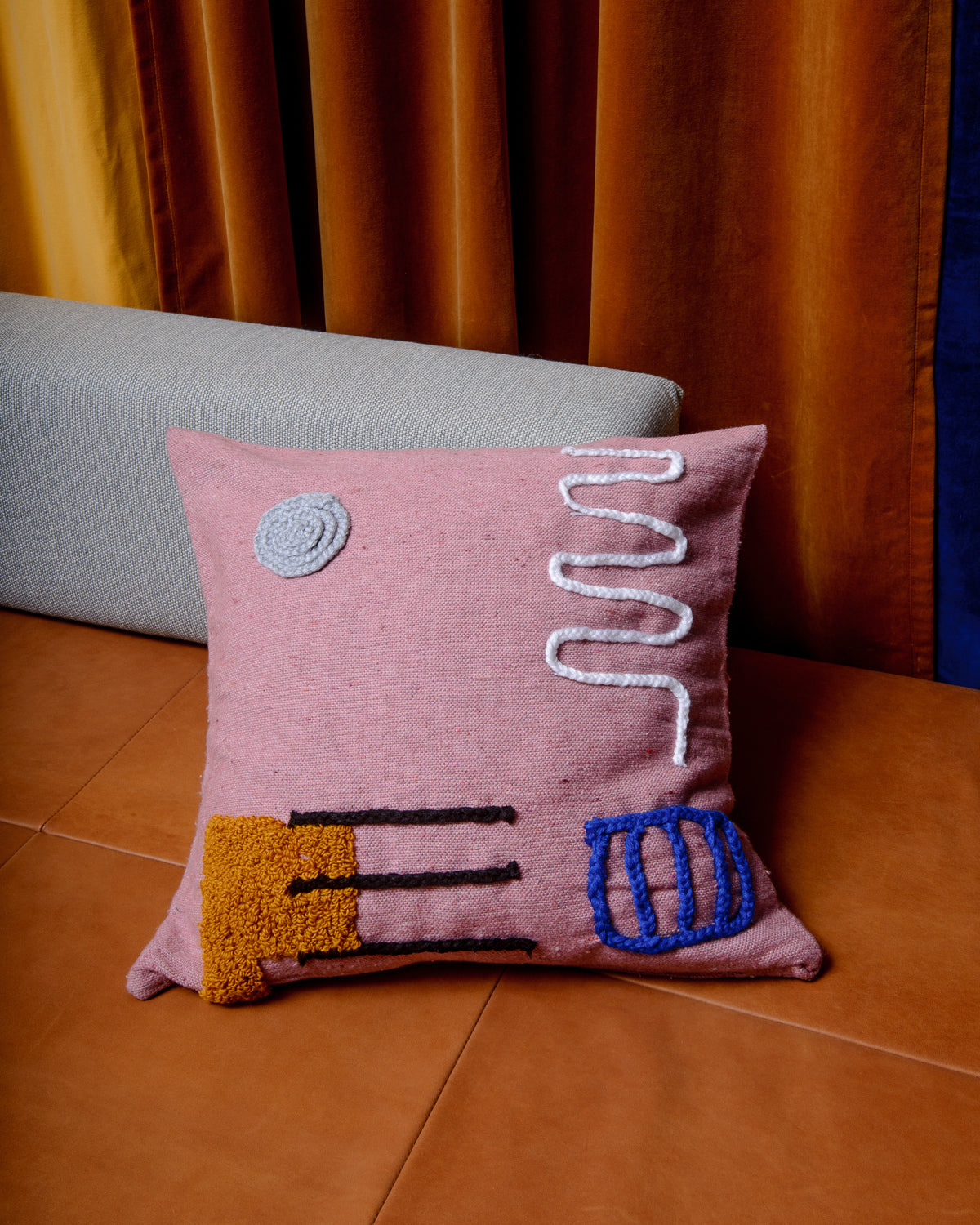 Pink cushion with hand embroidered abstract design on leather couch with velvet curtains behind.