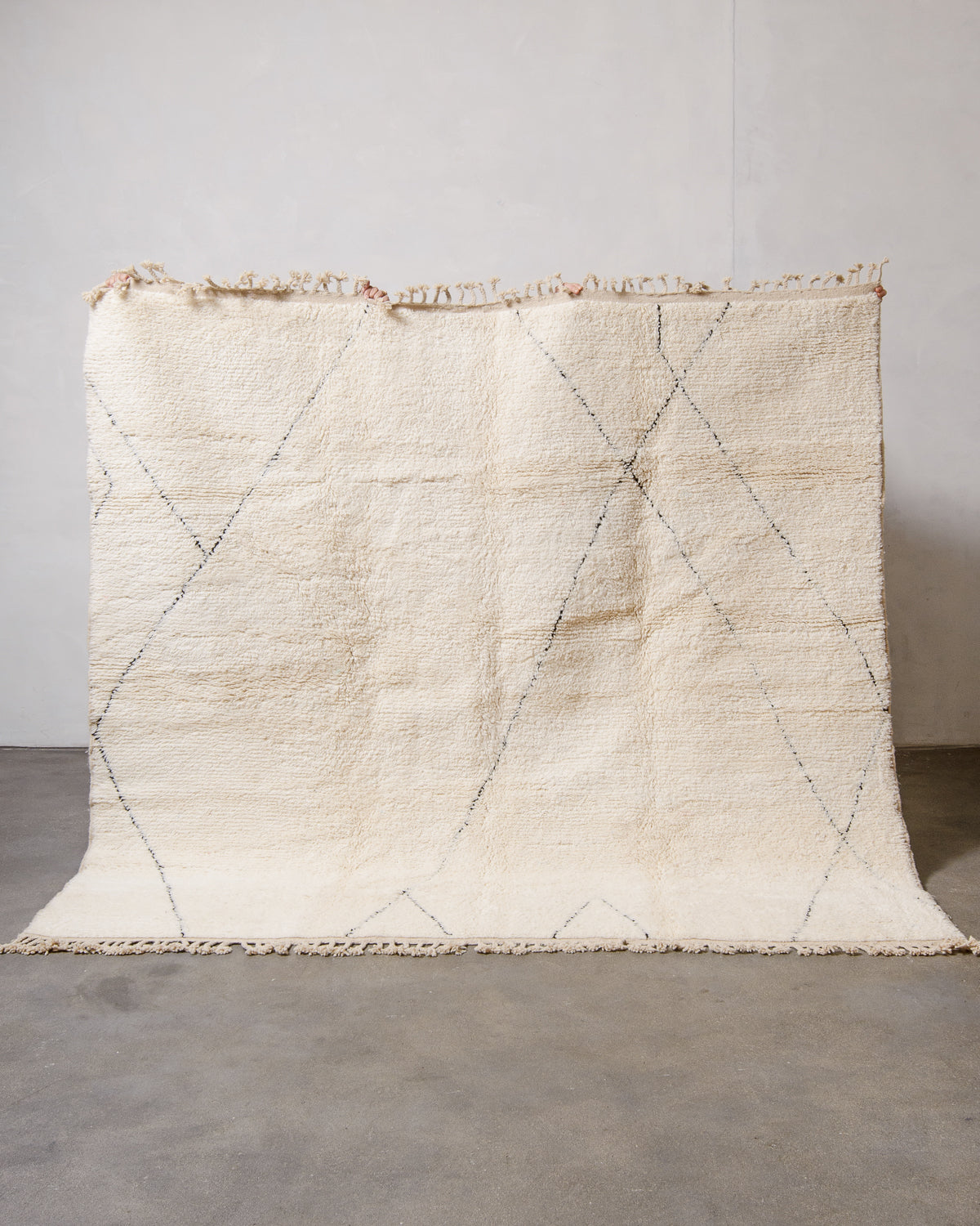 Cream wool Beniourain rug with minimal black line design being held up in front of a white wall and concrete floor. 