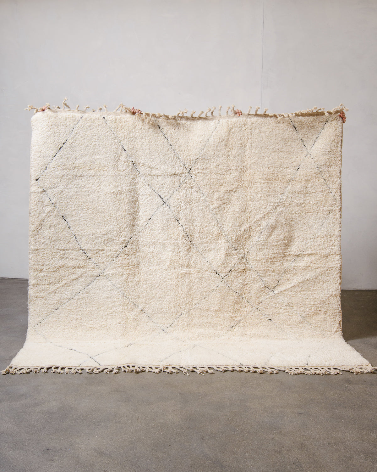 Cream wool Beniourain rug with minimal black line design being held up in front of a white wall and concrete floor. 