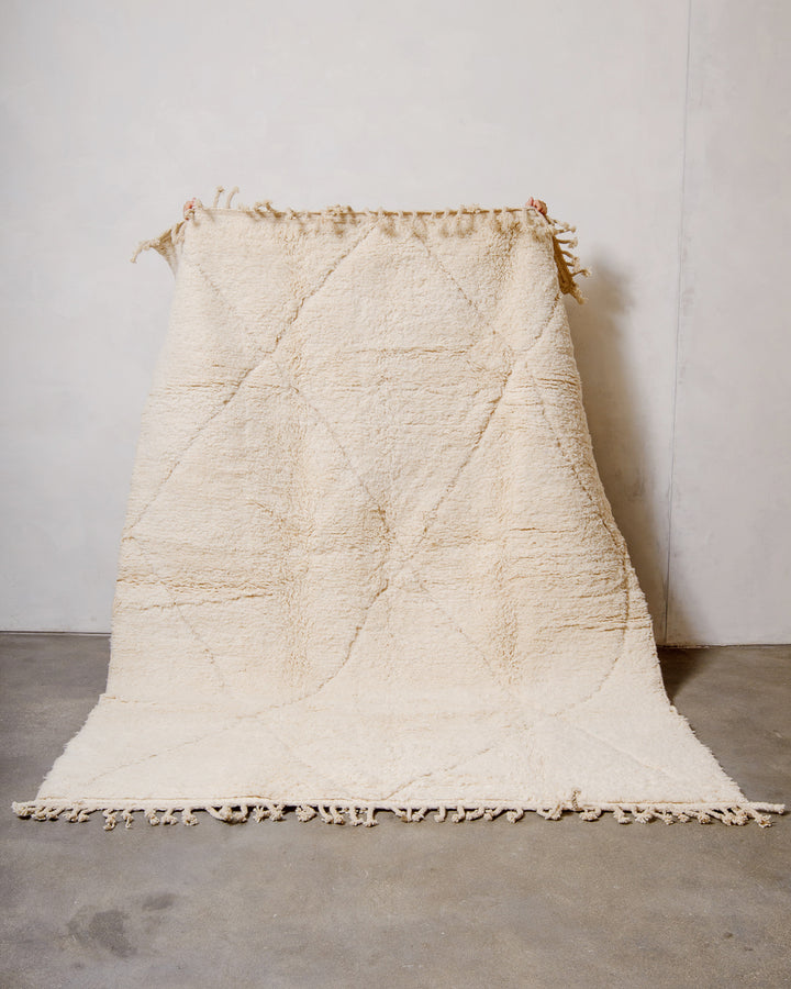 Wool Beniourain rug in natural cream with a subtle diamond design, being held up against a white wall. 