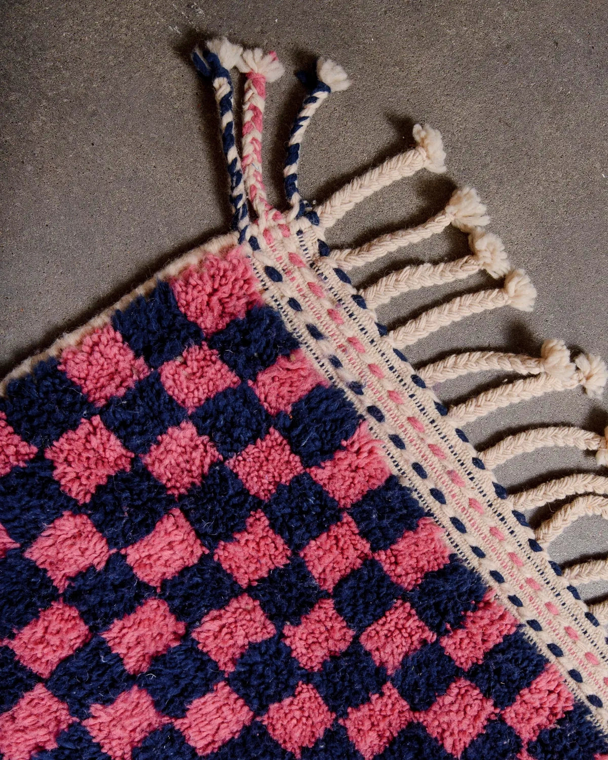 Tassels on corner of wool Berber rug with a navy and dark pink checkered pattern laying on concrete floor.