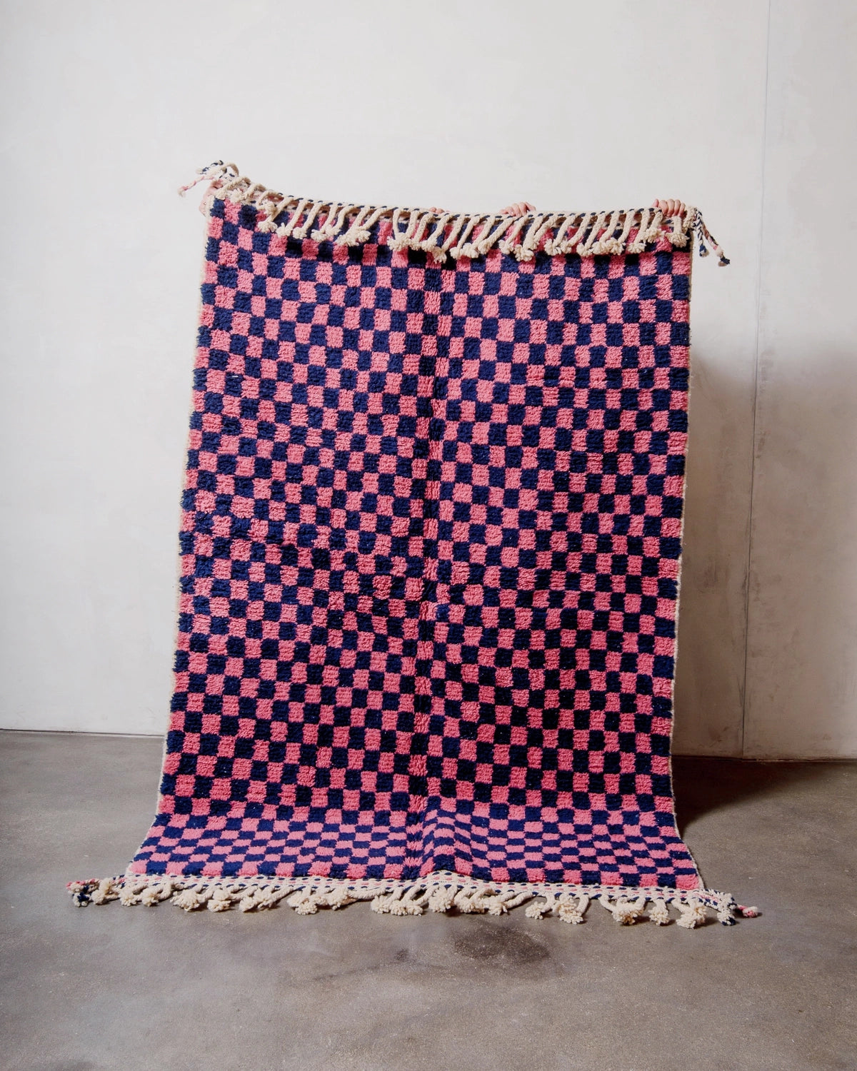 Berberlin Beniourain rug with a navy and dark pink checkered pattern being held up in front of a white wall. 