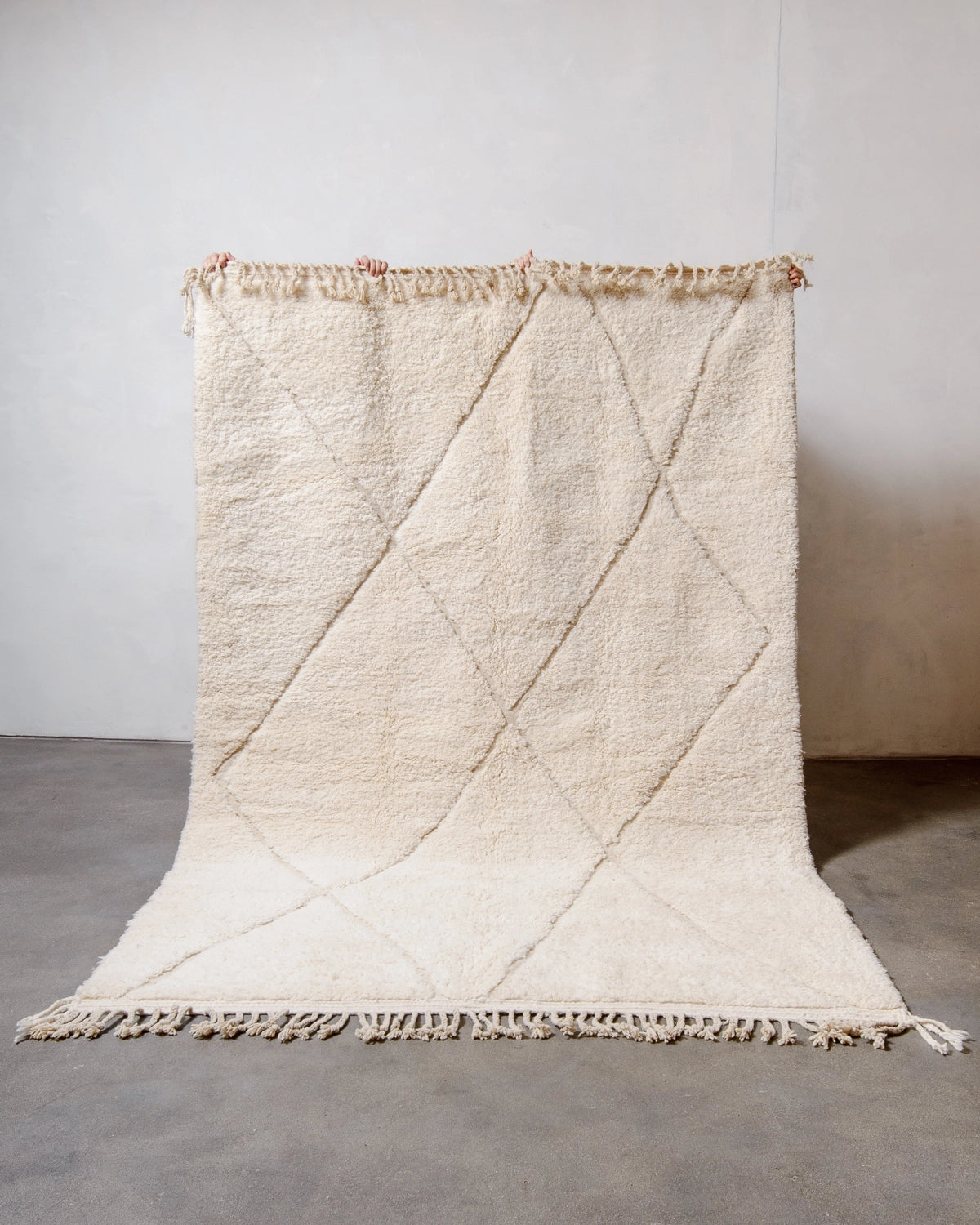 Berberlin Beniourain Rug made of natural cream wool with a woven diamond design, held up against a white wall and concrete floor.
