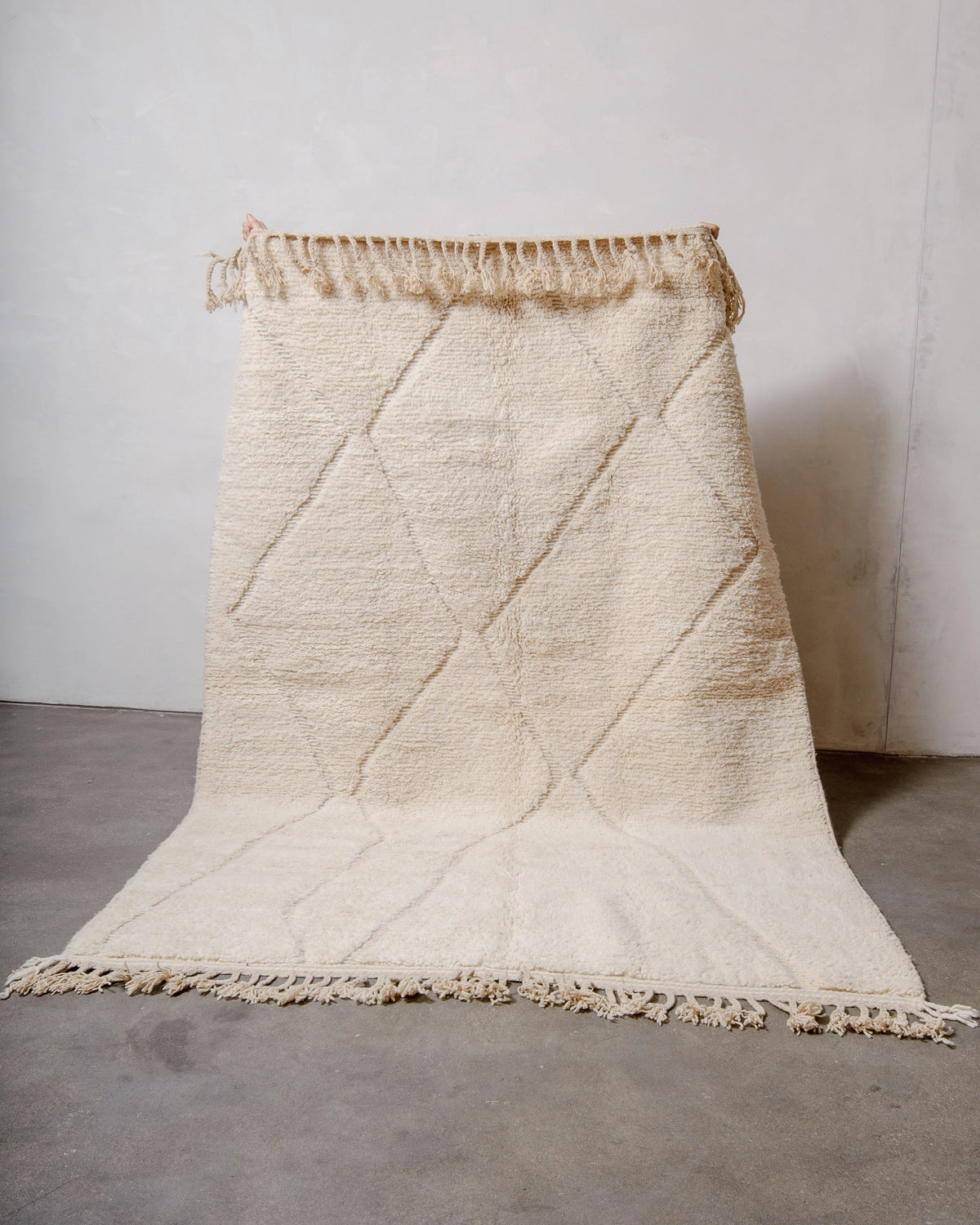 Berberlin Beniourain Rug made of natural cream wool with an abstract woven diamond design, held up against a white wall and concrete floor.