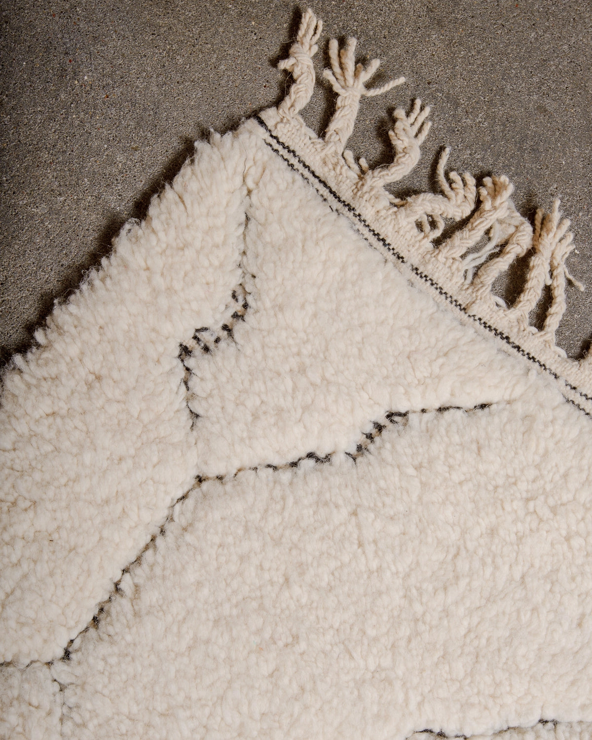 Natural wool tassels on a Berberlin Beniourain Rug made of cream wool with a subtle, abstract woven design in black, photographed on a concrete studio floor.
