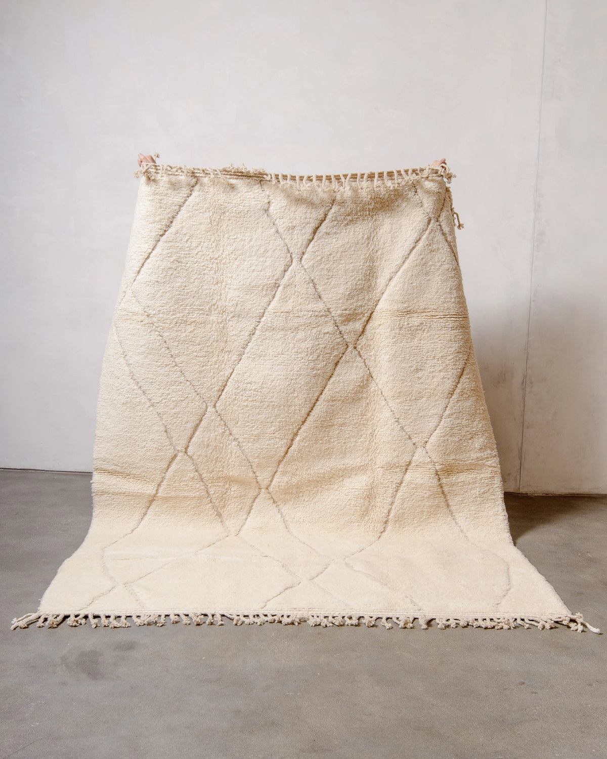 Berberlin Beniourain Rug made of natural cream wool with an abstract woven diamond design, held up against a white wall and concrete floor.