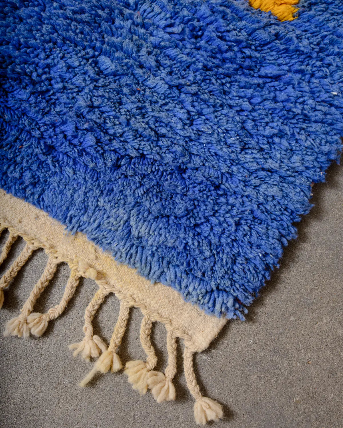 Tasseled edge and corner of handmade Berber featuring blue wool and laying on concrete floor. 