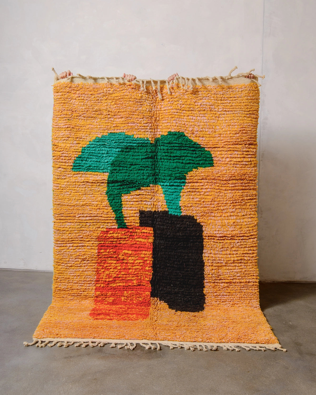 Berberlin vintage Berber rug made of wool in a background shade of mottled pink and orange, with overlapping forms in the centre in orange, black and green. The handwoven rug is being held up by four hands against a white wall.