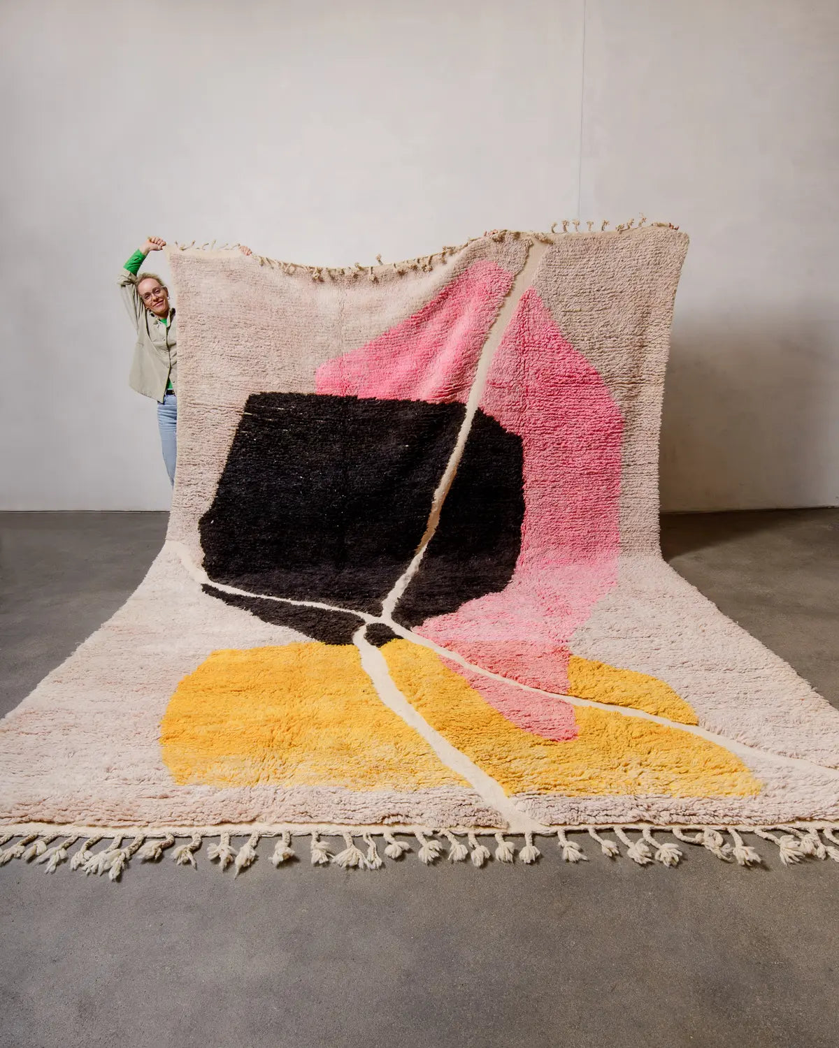 Large handcrafted Berberlin Berber rug from Morocco featuring shades of pink, yellow, black and cream being held up against white wall by a woman with green sleeves. 
