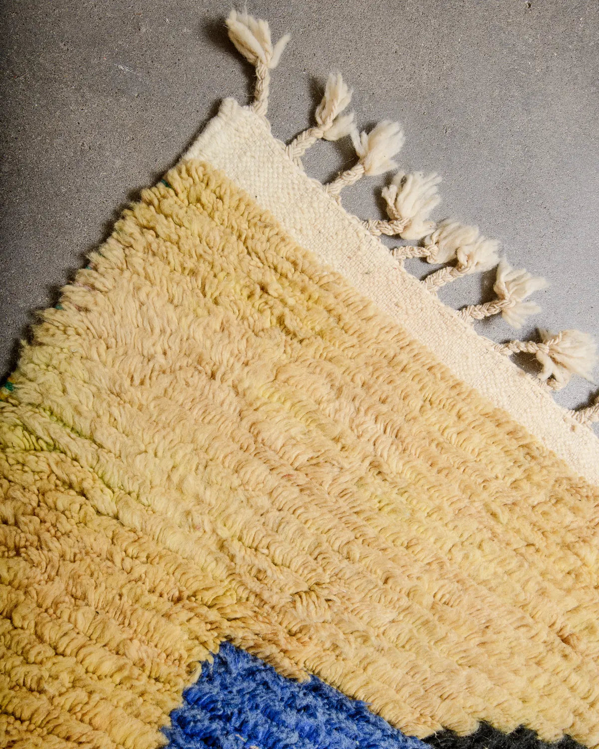 Cream tasseled edge of a Berberlin vintage Berber rug featuring yellow and blue wool, laying on concrete floor.
