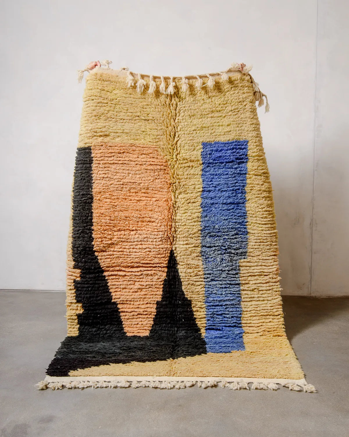 Handcrafted vintage Berber rug from Morocco in shades of yellow, apricot, blue and black, being held up against white wall and concrete floor. 