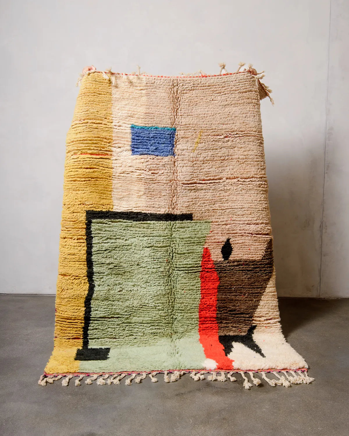 Berberlin vintage Berber rug in shades of sand, pistachio, red, yellow and blue in a modern, graphic design, being held up against a white wall. 