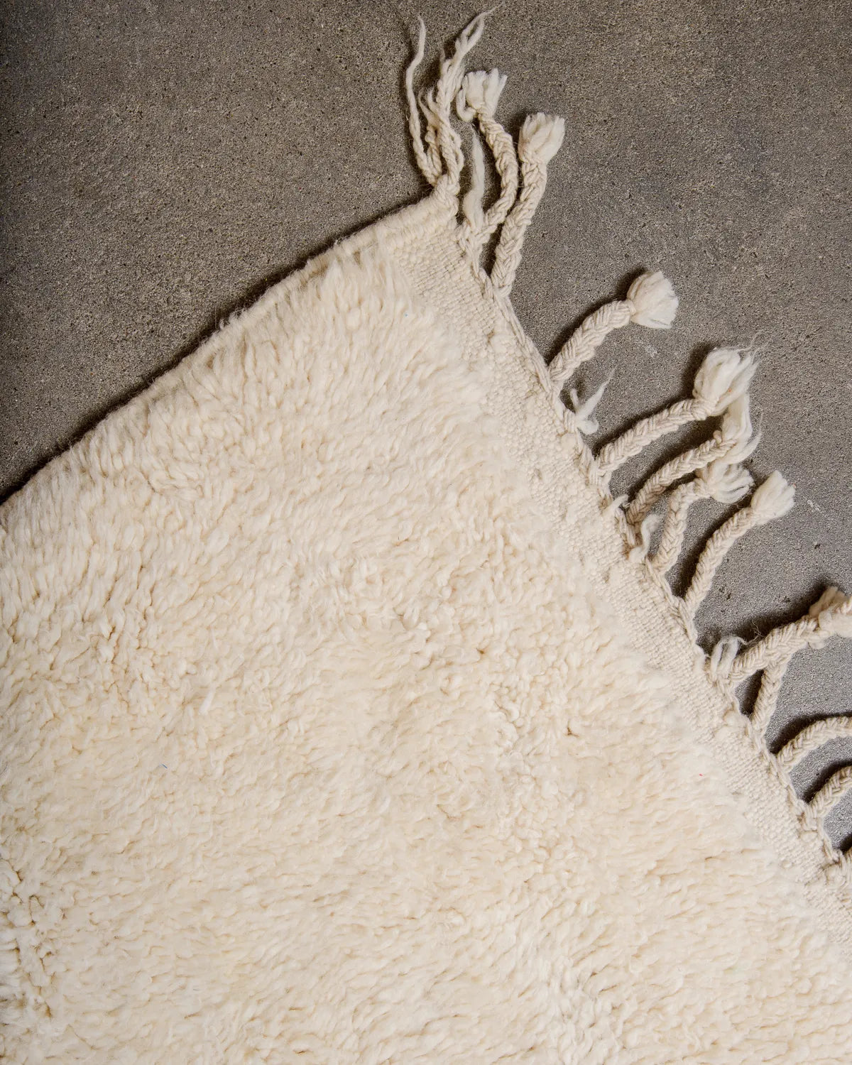 Tasseled edge of wool Berber rug in cream wool laying on concrete floor.