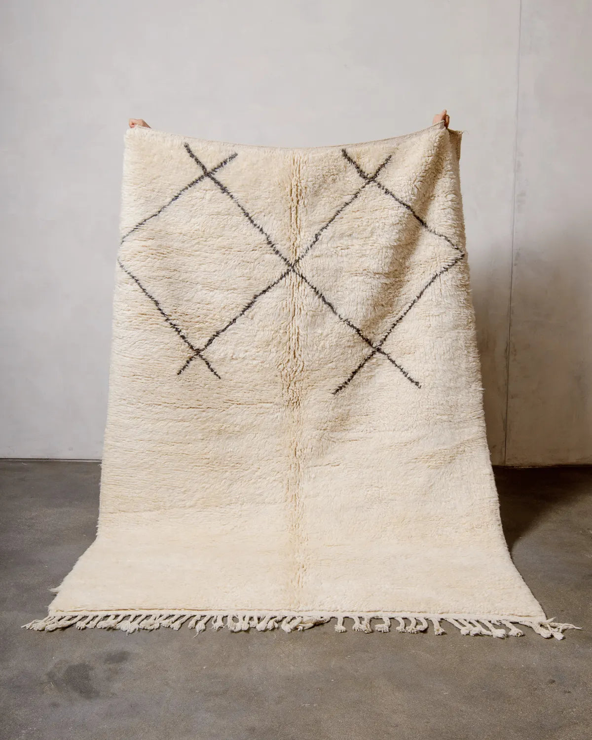 Berberlin vintage Berber rug in cream wool and minimal grey-brown line design, being held up against a white wall and concrete floor.