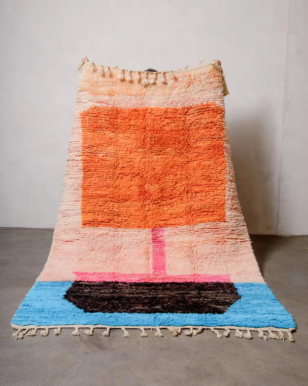 Berberlin vintage Berber rug in shades of peach, orange, pink, black and blue being held up against a white wall. 