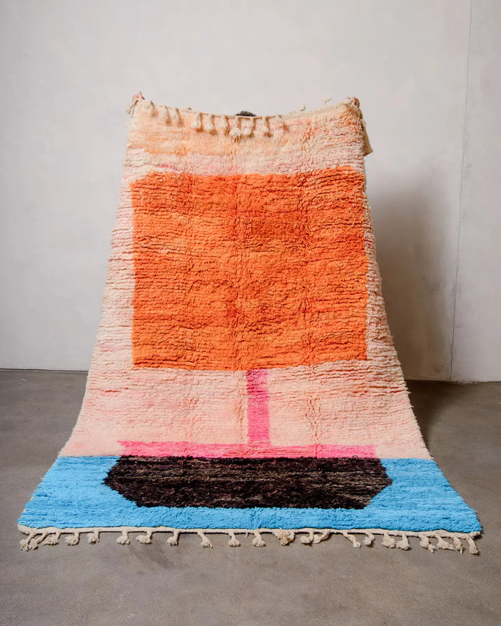 Berberlin vintage Berber rug in shades of peach, orange, pink, black and blue being held up against a white wall. 