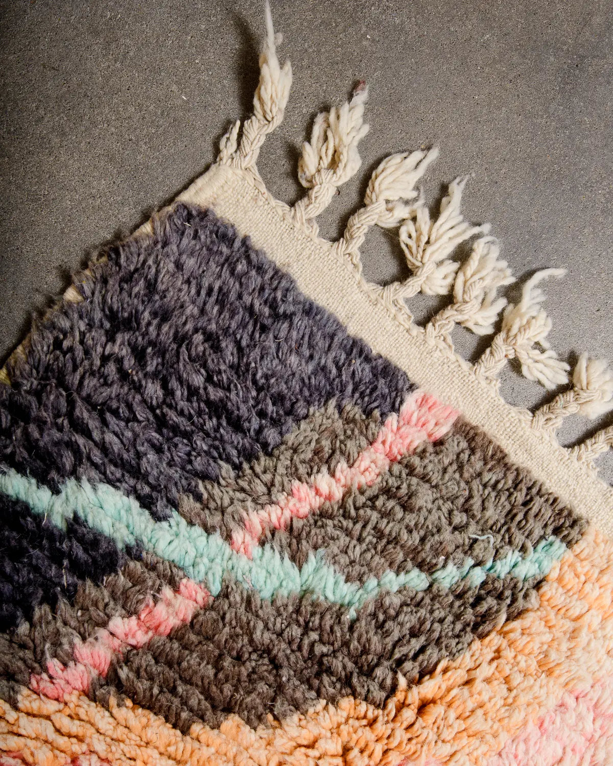 Tasseled corner of wool Berber rug in shades of cream, blue, pink, brown and orange laying on concrete floor. 