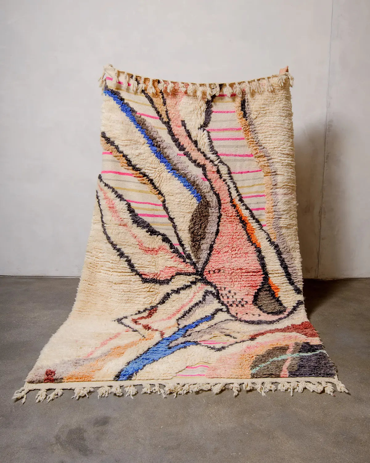 Berberlin Wool Berber rug in shades of cream, blue, pink and orange being held up against a white wall. The rug has a design made of knotted and flat-woven areas. 