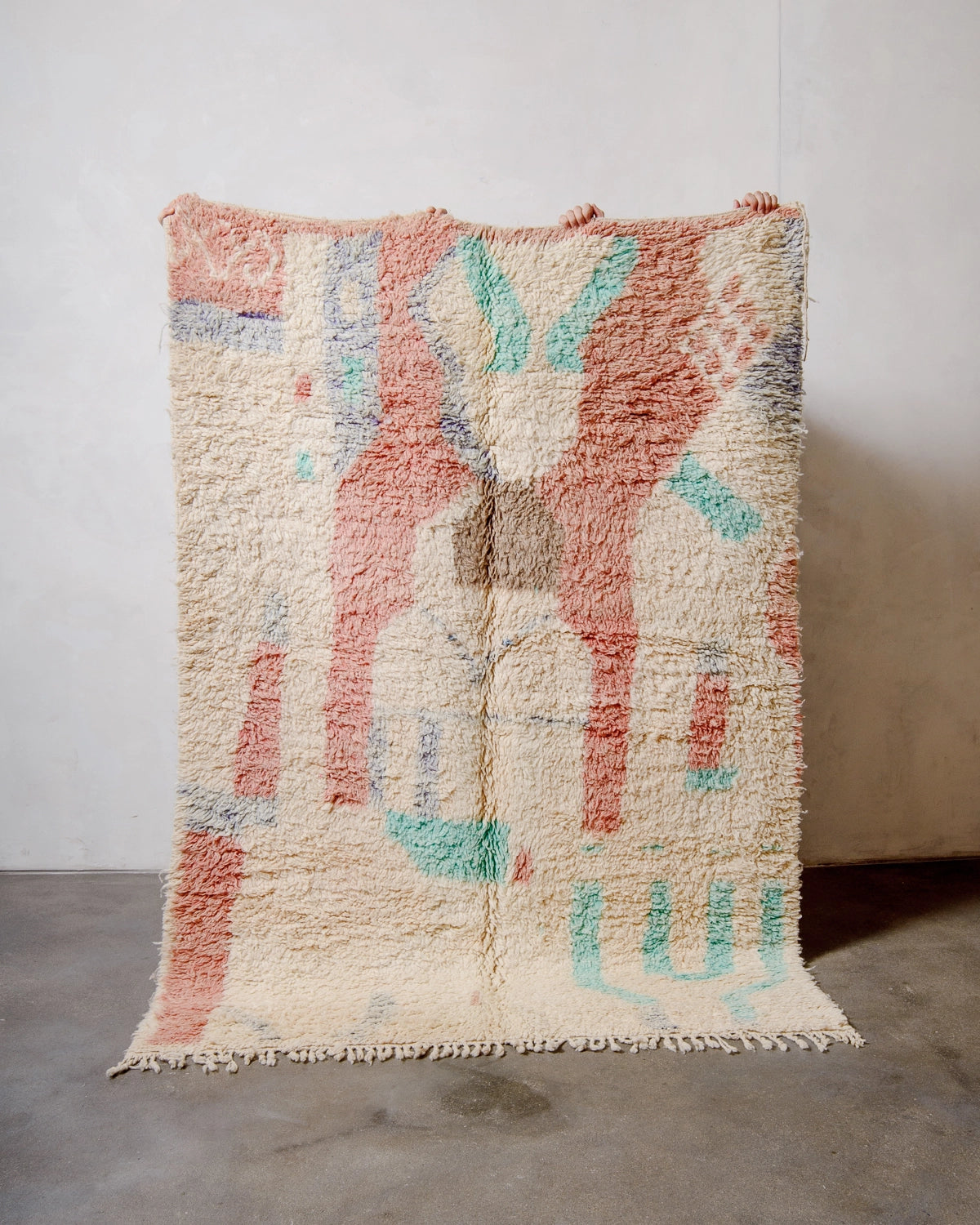 Berberlin Vintage Berber Rug made of fluffy cream, turquoise, faded red and purple wool, being held up against a white wall and grey concrete floor.