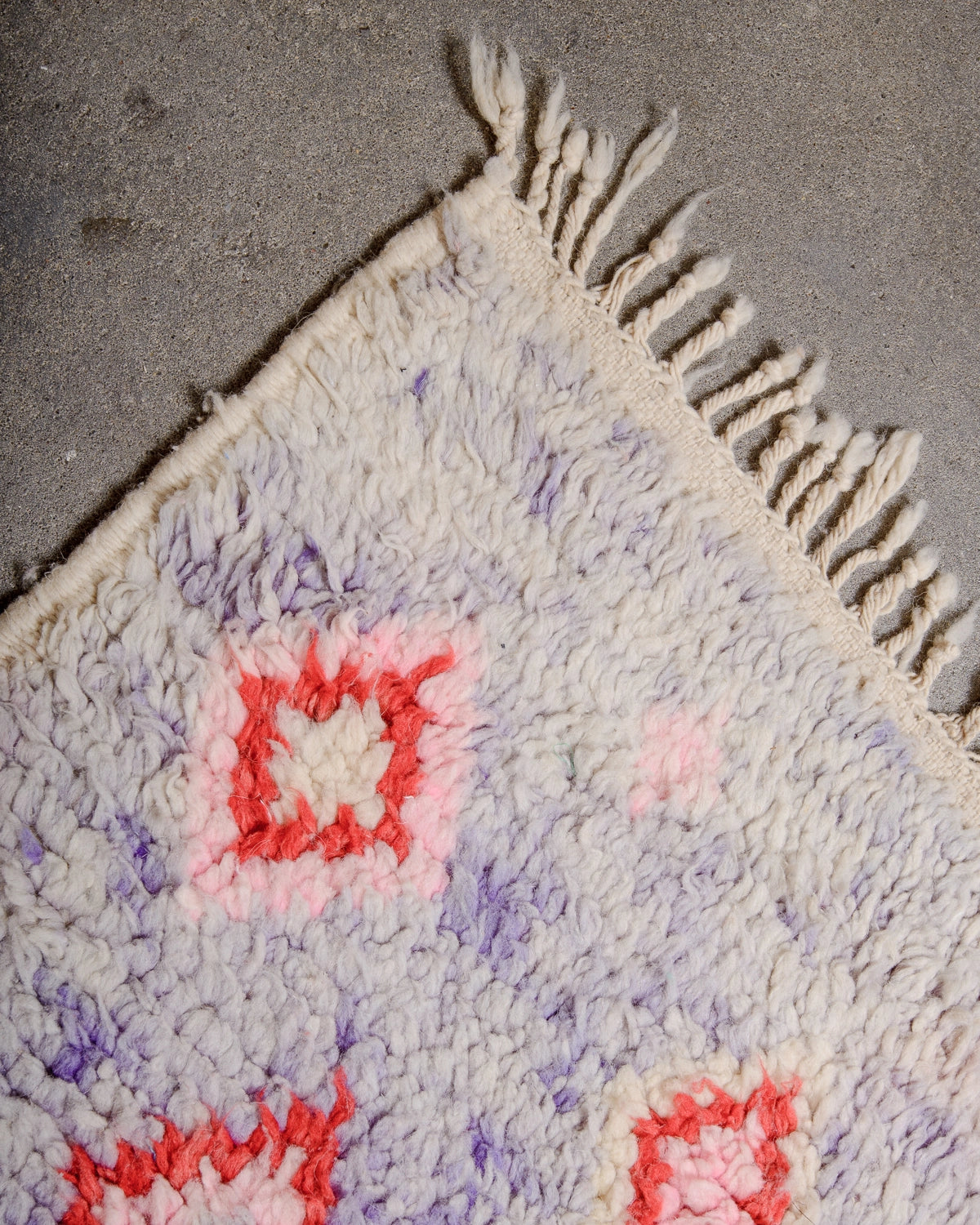 Natural wool tassels on a Berberlin Vintage Berber Rug made of fluffy lavender, red, pink and cream wool laying on concrete floor.