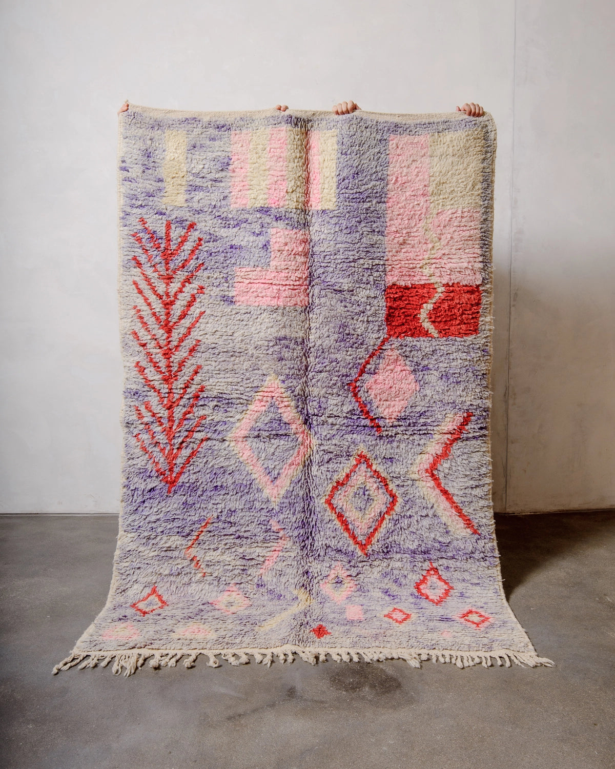 Berberlin Vintage Berber Rug made of fluffy lavender, red, pink and cream wool, being held up against a white wall and grey concrete floor.