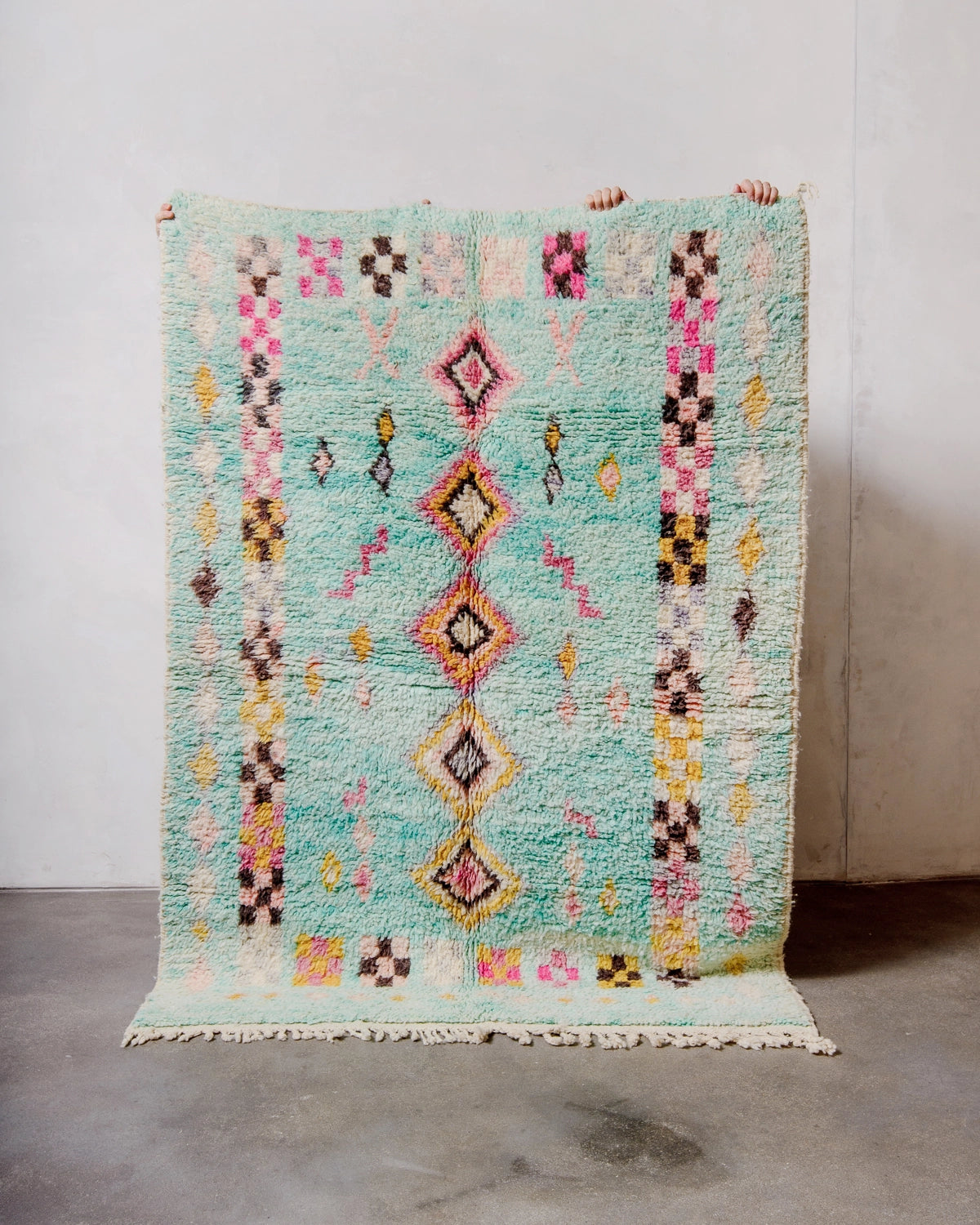 Berberlin Vintage Berber Rug made of fluffy turquoise wool, being held up against a white wall and grey concrete floor.