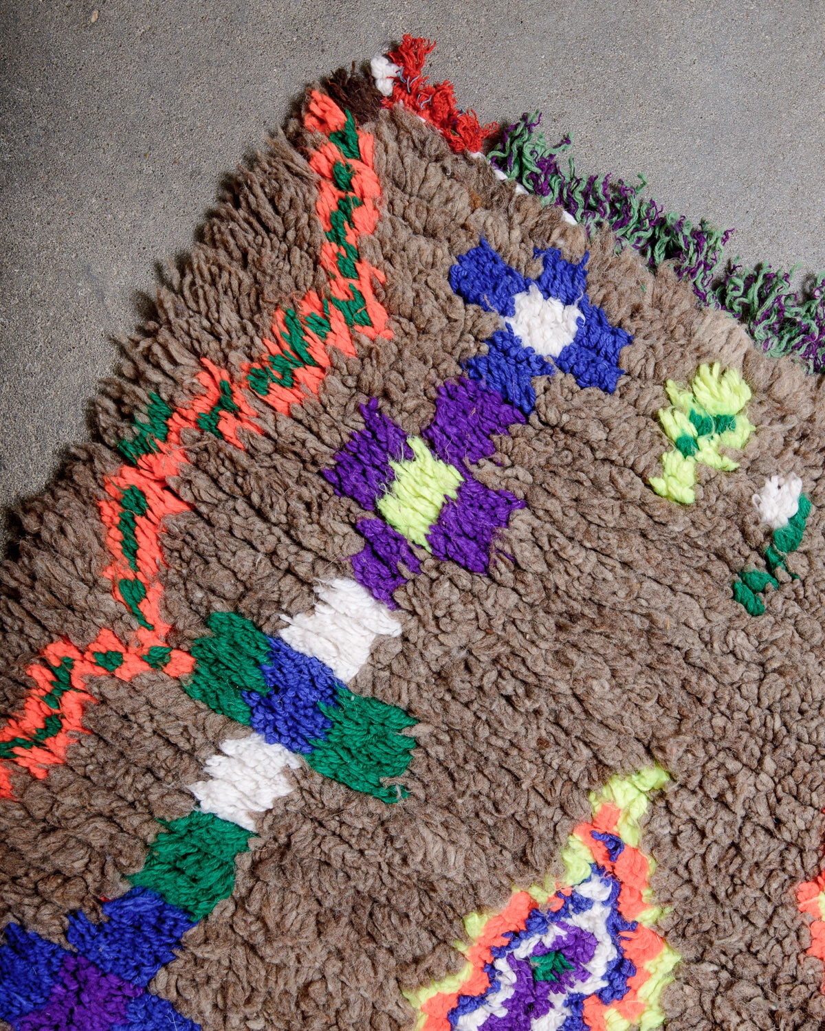 Detail of Vintage Berber Rug made of brown wool with design details in neon, purple, blue, green cotton laying on concrete.