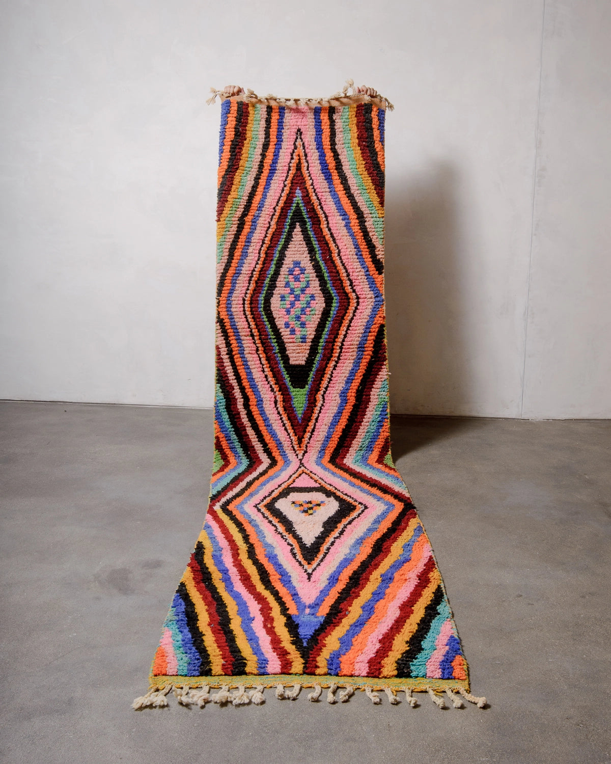 Berberlin Runner Rug made of brightly coloured wool and held up against a white wall and concrete floor. 