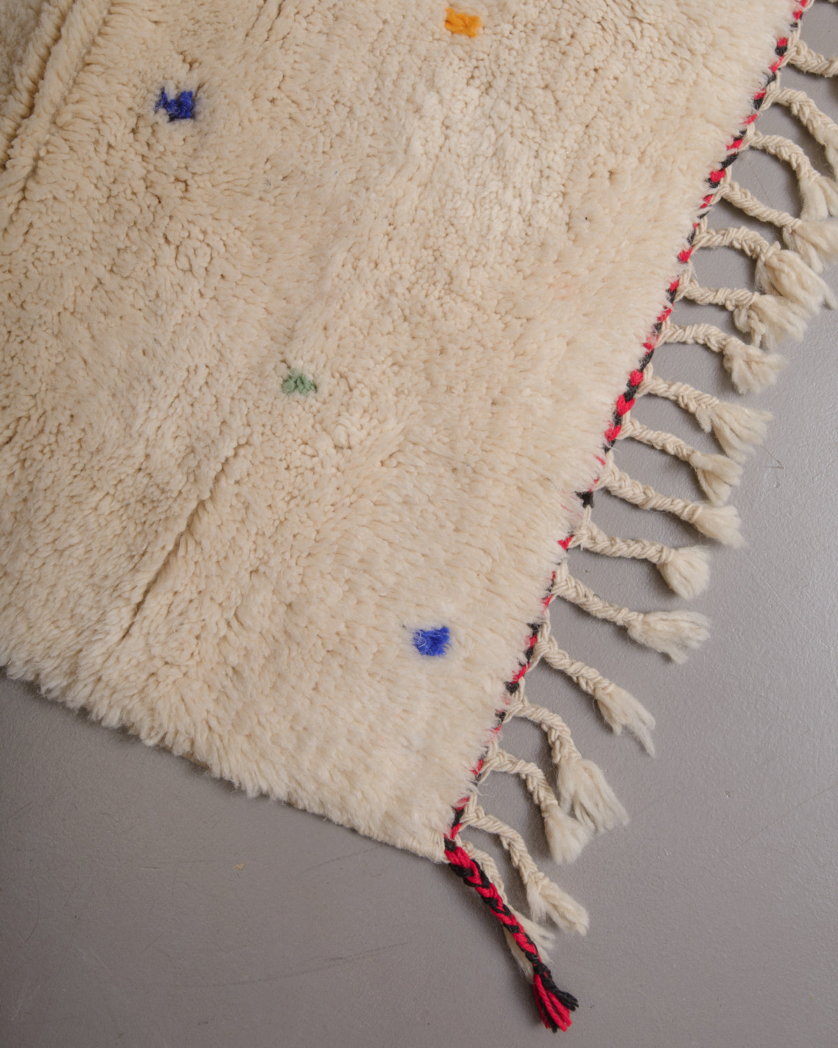 Corner of rug with cream wool and natural wool tassels and a black and red line across the edge.