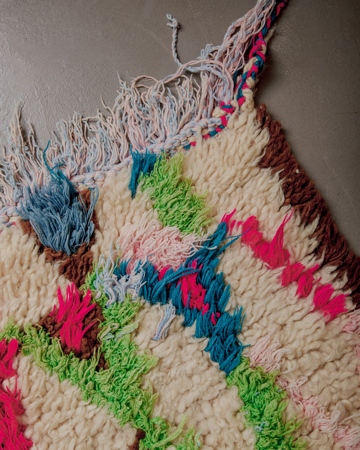 Corner of rug with green, pink and blue and a pink and blue cotton fringe.