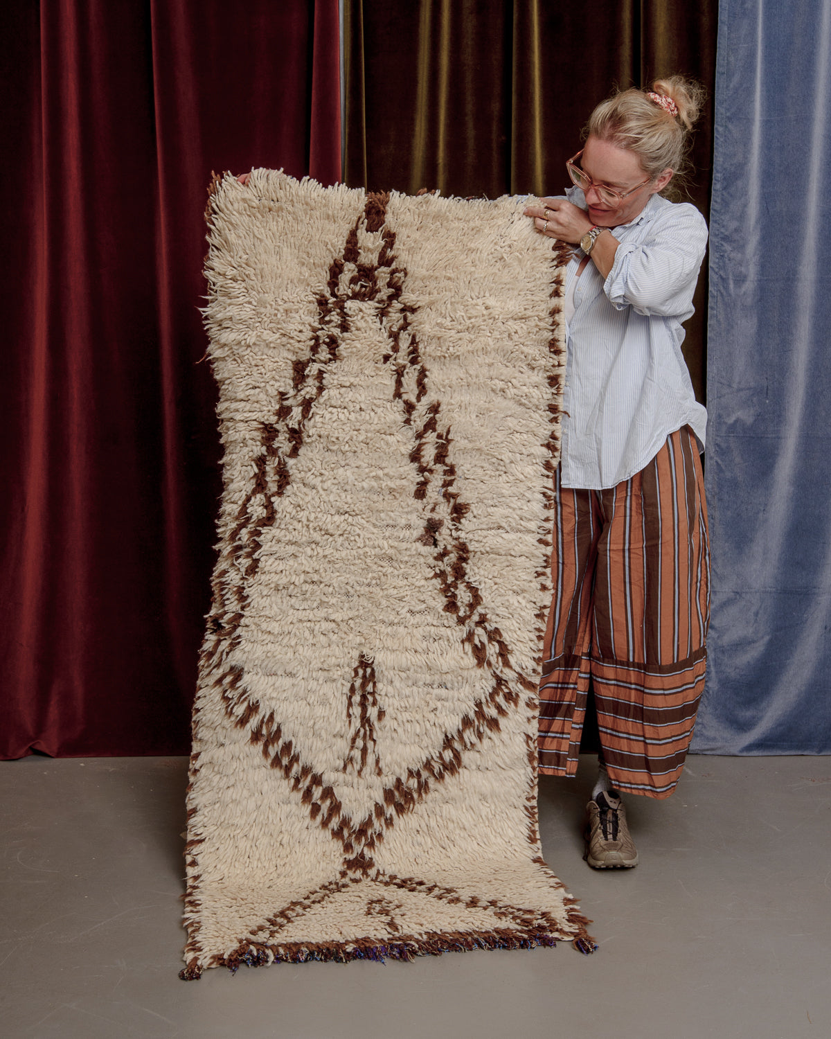Vintage Berberlin boucherouite runner rug being held up in front of a velvet curtain by a woman in striped pants.